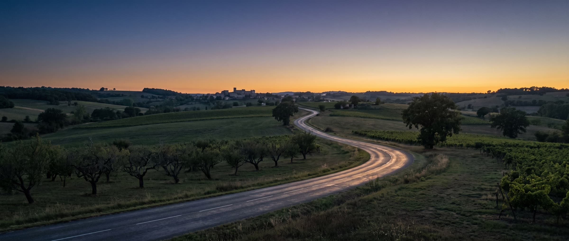 Une route de campagne serpente entre vergers et vignes au crépuscule, un village perché à l'horizon.